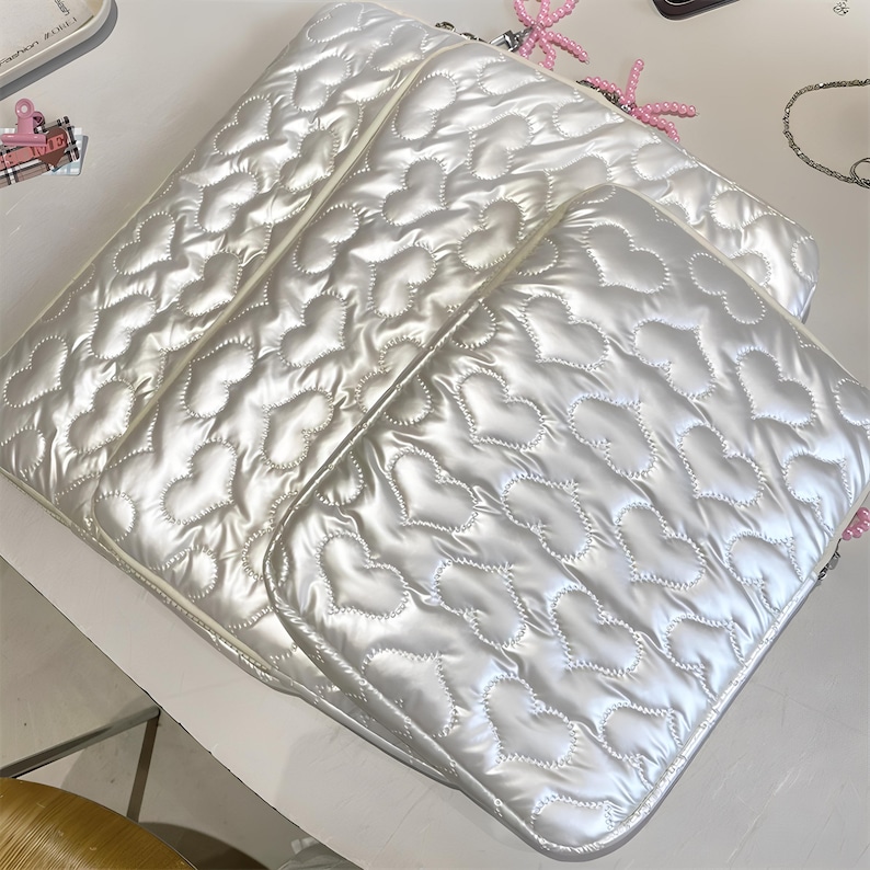 May include: Three white, quilted laptop sleeves with a heart pattern. The sleeves have a shiny, pearlescent finish and are stacked. The stitching creates a puffy, textured effect. The edges are neatly finished with a matching trim.