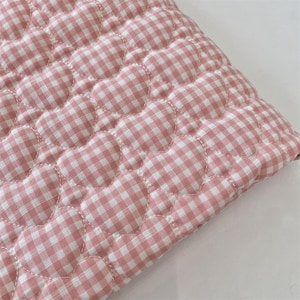May include: A quilted fabric textile featuring a repeating pattern of stitched hearts. The material displays a pink and white gingham check design, with the heart shapes defined by white thread stitching across the soft, padded surface.