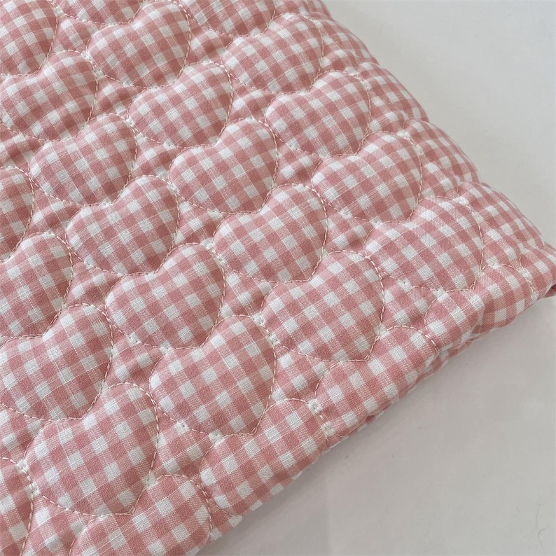 May include: A quilted fabric textile featuring a repeating pattern of stitched hearts. The material displays a pink and white gingham check design, with the heart shapes defined by white thread stitching across the soft, padded surface.
