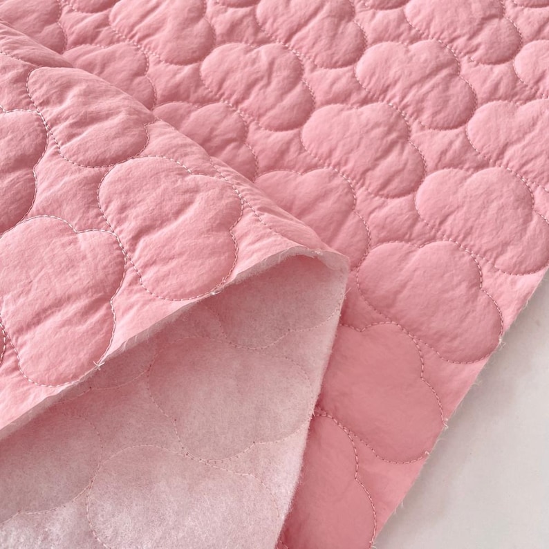 Op de afbeelding: Een stuk lichtroze doorgestikte stof met een herhalend wolkvormig stikpatroon. Het materiaal heeft een gladde, matte toplaag en een zachte, pluizige, lichtgekleurde onderkant die zichtbaar is waar de stof over zichzelf is gevouwen.