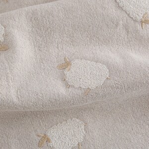 Puede incluir: Primer plano de una tela beige con ovejas bordadas en blanco. La tela tiene una textura sutil y un aspecto suave y natural.