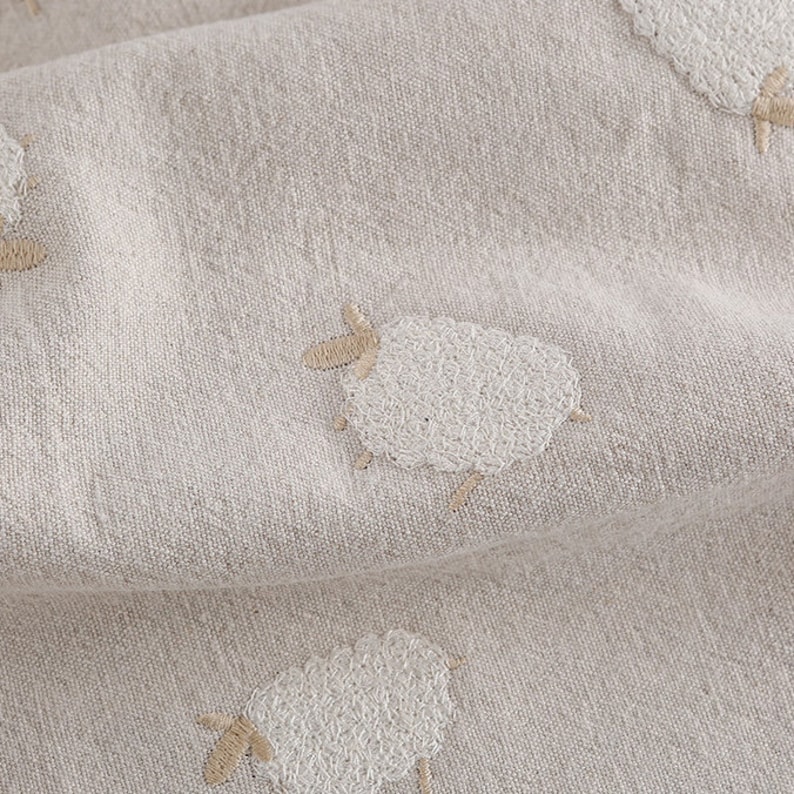 Puede incluir: Primer plano de una tela beige con ovejas bordadas en blanco. La tela tiene una textura sutil y un aspecto suave y natural.