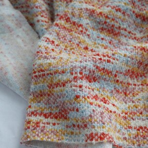 May include: A textured knit fabric featuring a variegated pattern of horizontal stripes in shades of bright orange, soft yellow, and pale blue against a cream-colored base. The material has a visible, slightly raised weave with a soft appearance.