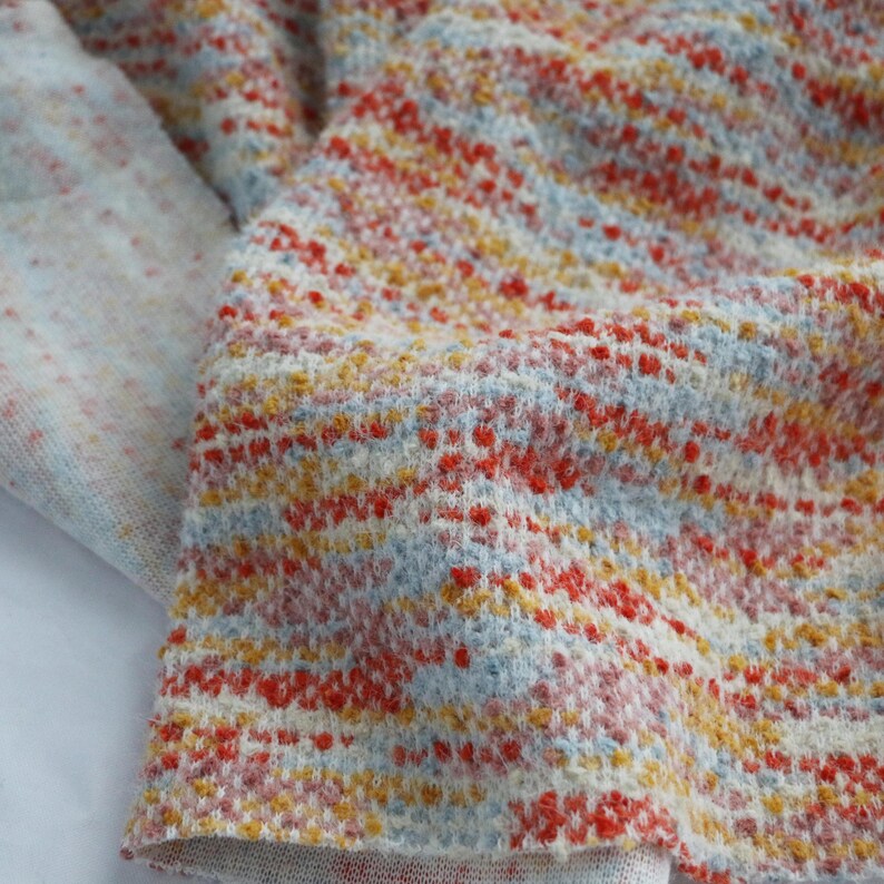 May include: A textured knit fabric featuring a variegated pattern of horizontal stripes in shades of bright orange, soft yellow, and pale blue against a cream-colored base. The material has a visible, slightly raised weave with a soft appearance.