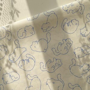May include: Off-white fabric with a repeating pattern of blue cat outlines in various poses. The cats are drawn in a simple, playful style, with details like whiskers and curved tails. The fabric is slightly wrinkled, suggesting it's a soft material.