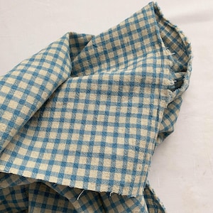 May include: Close-up of a folded piece of fabric with a blue and beige gingham pattern. The fabric is textured and appears to be cotton or linen. The edges of the fabric are frayed, suggesting a handmade or vintage item.