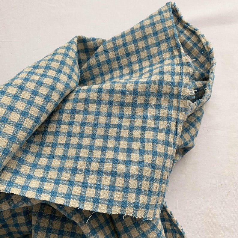 May include: Close-up of a folded piece of fabric with a blue and beige gingham pattern. The fabric is textured and appears to be cotton or linen. The edges of the fabric are frayed, suggesting a handmade or vintage item.