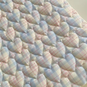 May include: Close-up of a quilted fabric with a repeating heart pattern. The hearts are filled with a plaid design in shades of pink, blue, and white. The hearts are stitched onto a white background.