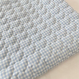 May include: A light blue and white gingham fabric featuring a repeating quilted heart pattern. The material has a soft, padded texture with visible stitching outlining each individual heart shape across the entire surface of the textile.