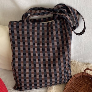 May include: A large tote bag with a checkered pattern in shades of brown, black, and blue. The bag has two long handles for carrying over the shoulder or by hand. The bag is made of a sturdy fabric.