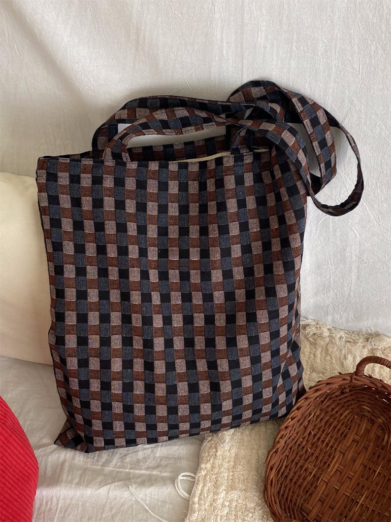 May include: A large tote bag with a checkered pattern in shades of brown, black, and blue. The bag has two long handles for carrying over the shoulder or by hand. The bag is made of a sturdy fabric.
