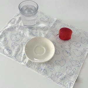 May include: A white placemat with a blue cat pattern, a clear glass of water, a white saucer, and a red apple. The placemat is rectangular and has a stitched edge. The cat illustrations are simple line drawings.