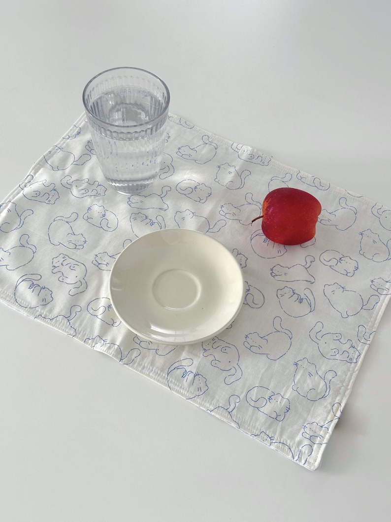 May include: A white placemat with a blue cat pattern, a clear glass of water, a white saucer, and a red apple. The placemat is rectangular and has a stitched edge. The cat illustrations are simple line drawings.