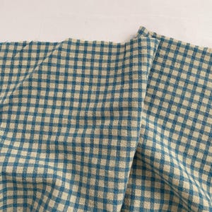 Pode incluir: Uma imagem em close-up de um tecido dobrado com um padrão xadrez vichy azul e bege. O tecido é texturizado e tem uma aparência ligeiramente enrugada. O padrão consiste em quadrados espaçados uniformemente.