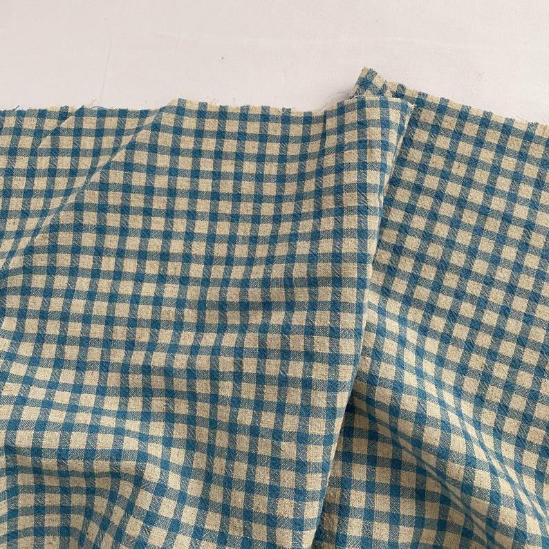 May include: A close-up of a folded fabric with a blue and beige gingham pattern. The fabric is textured and has a slightly wrinkled appearance. The pattern consists of evenly spaced squares.