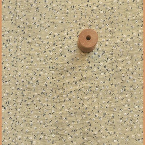 May include: A light tan fabric with a small floral pattern of white flowers and dark blue leaves. A wooden spool with a central hole sits on the fabric. The fabric has a textured appearance.