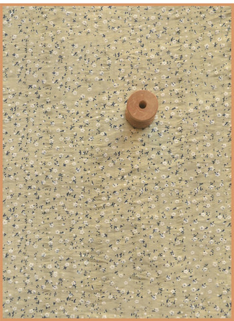 May include: A light tan fabric with a small floral pattern of white flowers and dark blue leaves. A wooden spool with a central hole sits on the fabric. The fabric has a textured appearance.