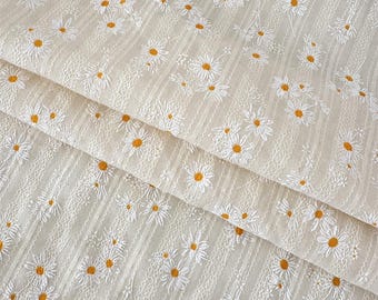 Op de afbeelding: Een lichtbeige stof met een herhalend patroon van geborduurde witte madeliefjes met gele harten. Het textiel bevat verticale gestructureerde strepen en fijn stiksel tussen de bloemmotieven, wat zorgt voor een gelaagd uiterlijk.