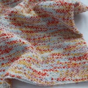 May include: A textured fabric featuring a woven pattern of cream, light blue, bright orange, and yellow threads. The material has a soft, fuzzy appearance with a multi-colored speckled effect created by the interlacing of these distinct yarn colors.
