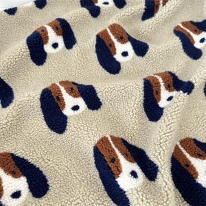 Peut inclure: Un tissu en polaire sherpa beige avec un motif répétitif de têtes de chien. Chaque tête de chien est représentée en blanc, marron et bleu marine foncé, avec de longues oreilles bleu marine, une tache marron sur le front et un museau blanc.