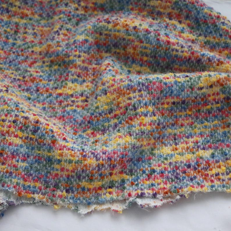 May include: A textured fabric featuring a multi-colored speckled pattern with vibrant hues of yellow, blue, red, and purple. The material has a loose, woven appearance with a soft, fuzzy surface and an uneven, frayed edge along the bottom border.