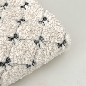 Peut inclure: Gros plan sur un tissu crème texturé avec un motif répété de nœuds noirs. Le tissu a un aspect doux et moelleux, avec les nœuds cousus en quadrillage.