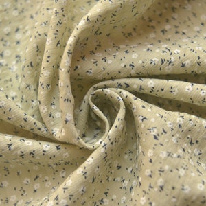 May include: Close-up of a light green fabric with a delicate floral pattern. The fabric is covered in tiny white flowers and small dark blue accents, creating a soft, textured appearance. The fabric is gathered in soft folds.
