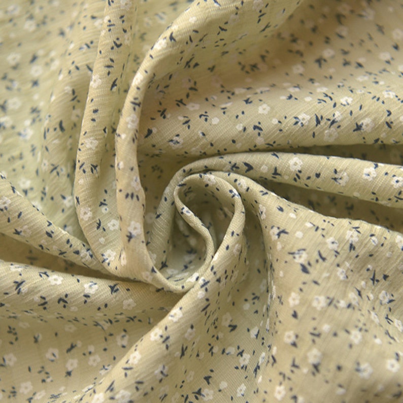 May include: Close-up of a light green fabric with a delicate floral pattern. The fabric is covered in tiny white flowers and small dark blue accents, creating a soft, textured appearance. The fabric is gathered in soft folds.
