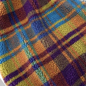 Könnte beinhalten: Ein strukturierter Sherpa-Fleece-Stoff mit einem mehrfarbigen Karomuster. Das Design besteht aus sich kreuzenden Linien und Blöcken in leuchtenden Tönen von Orange, Violett, leuchtendem Blau und Limettengrün, was eine dichte, flauschige und weiche Oberfläche ergibt.