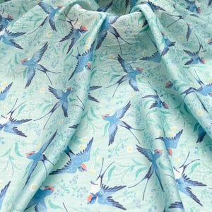 May include: A light blue fabric with a repeating pattern of blue swallows and small white flowers. The swallows have red accents on their heads and are depicted in flight. The fabric has a slightly wrinkled texture.