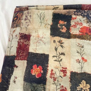 Puede incluir: Tela doblada con un diseño de patchwork que presenta patrones florales en tonos rojos, naranjas y beige. La tela tiene un aspecto vintage y desgastado, con una mezcla de bloques cuadrados y rectangulares.