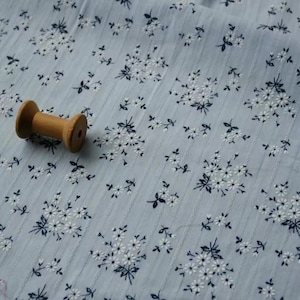 K&ouml;nnte beinhalten: Nahaufnahme eines hellblauen Stoffes mit einem wei&szlig;en und schwarzen Blumenmuster. Eine Holzspule mit Garn liegt auf dem Stoff.