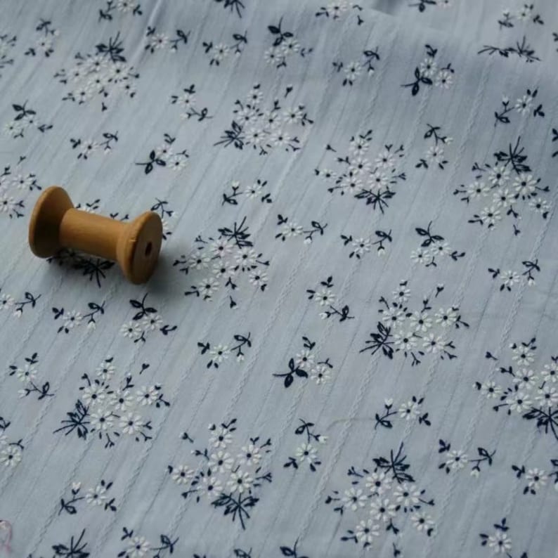 K&ouml;nnte beinhalten: Nahaufnahme eines hellblauen Stoffes mit einem wei&szlig;en und schwarzen Blumenmuster. Eine Holzspule mit Garn liegt auf dem Stoff.