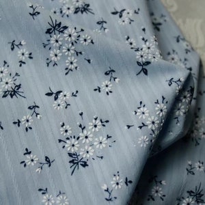 K&ouml;nnte beinhalten: Ein hellblauer Stoff mit einem wei&szlig;en Blumenmuster. Der Stoff hat eine subtile gestreifte Textur.