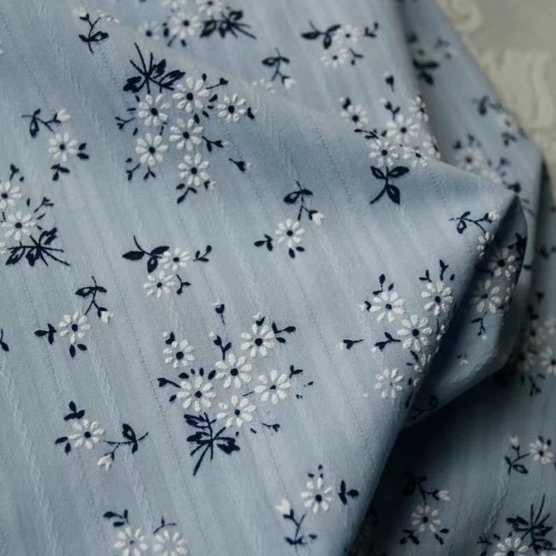 K&ouml;nnte beinhalten: Ein hellblauer Stoff mit einem wei&szlig;en Blumenmuster. Der Stoff hat eine subtile gestreifte Textur.
