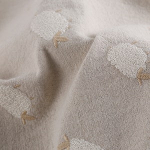 Puede incluir: Primer plano de una tela beige con dise&ntilde;os de ovejas bordadas en blanco. La tela tiene una apariencia texturizada, similar al lino.