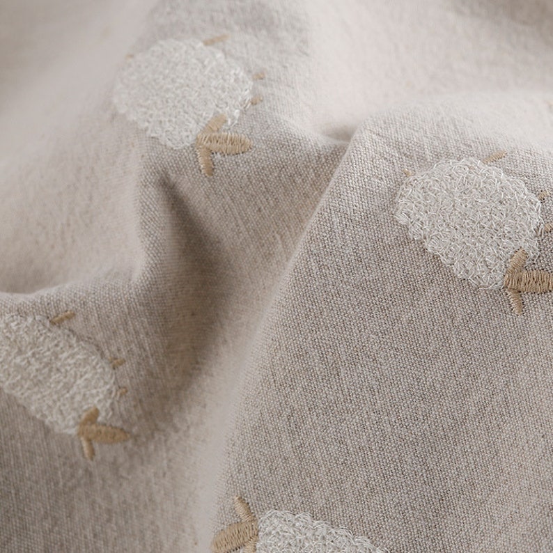 Puede incluir: Primer plano de una tela beige con dise&ntilde;os de ovejas bordadas en blanco. La tela tiene una apariencia texturizada, similar al lino.
