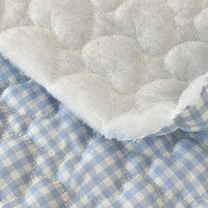 May include: A quilted fabric featuring a light blue and white gingham check pattern. The material is stitched with a repeating heart-shaped design, creating a textured, puffy surface. A corner of the fabric is folded over to reveal a white, fuzzy lining.