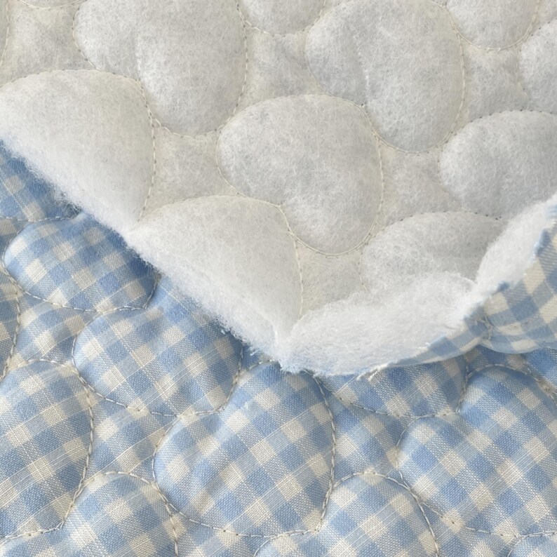 May include: A quilted fabric featuring a light blue and white gingham check pattern. The material is stitched with a repeating heart-shaped design, creating a textured, puffy surface. A corner of the fabric is folded over to reveal a white, fuzzy lining.