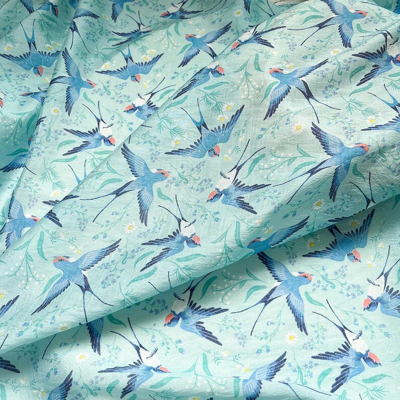 May include: A light blue fabric with a repeating pattern of blue swallows in flight. The swallows have red accents on their heads and are set against a background of white flowers and green foliage.