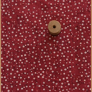 May include: A wooden spool sits on a red fabric with a white floral pattern. The fabric has a textured appearance, and the flowers are small and evenly spaced across the surface. The spool is light brown and has a hole in the center.