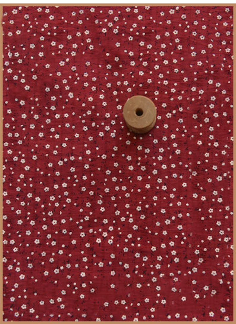 May include: A wooden spool sits on a red fabric with a white floral pattern. The fabric has a textured appearance, and the flowers are small and evenly spaced across the surface. The spool is light brown and has a hole in the center.