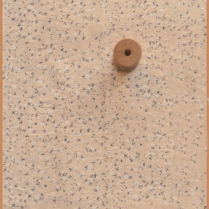 May include: A small, round, wooden spool with a central hole rests on a beige fabric patterned with tiny white and dark blue floral designs. The fabric is framed by a thin, brown border.
