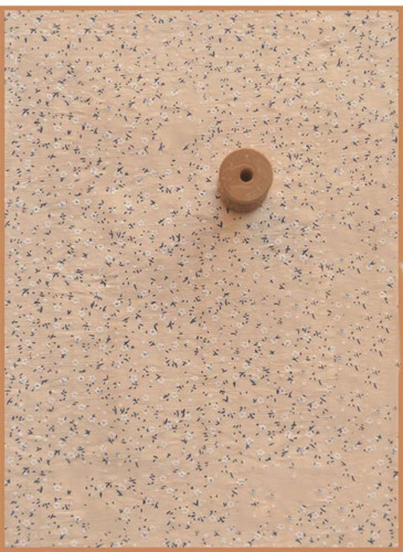 May include: A small, round, wooden spool with a central hole rests on a beige fabric patterned with tiny white and dark blue floral designs. The fabric is framed by a thin, brown border.