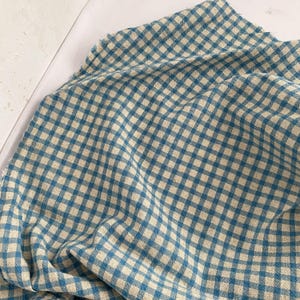 May include: A close-up of a blue and beige checkered fabric. The fabric is folded and draped, showing the pattern of small squares. The fabric appears to be a soft, woven material, possibly cotton or linen. The color scheme is a classic, timeless design.