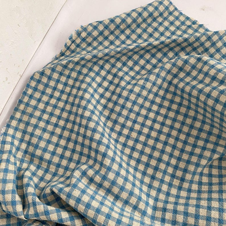 May include: A close-up of a blue and beige checkered fabric. The fabric is folded and draped, showing the pattern of small squares. The fabric appears to be a soft, woven material, possibly cotton or linen. The color scheme is a classic, timeless design.