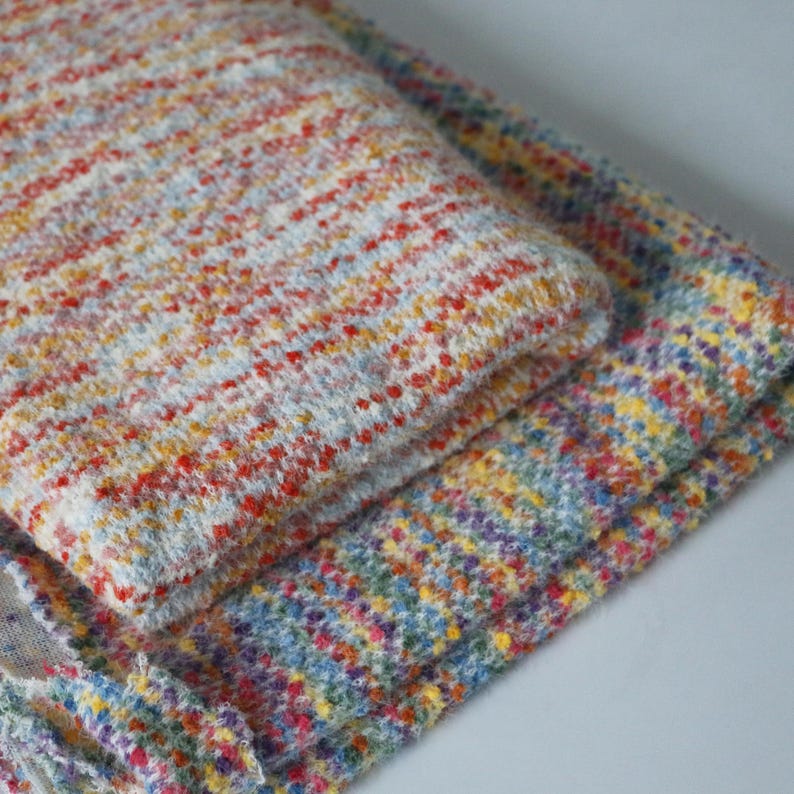 May include: A folded piece of fabric featuring a textured, multi-colored knit pattern. The material displays a vibrant mix of red, yellow, blue, and purple flecks woven into a white base, creating a speckled appearance across the entire surface.