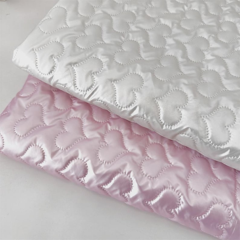 May include: Two pieces of quilted fabric, one white and one pink, are layered. Both feature a heart-shaped pattern stitched across the surface. The fabric has a shiny, satin-like appearance, reflecting light.