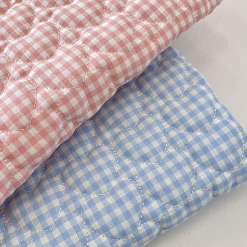 May include: Two quilted fabric pieces featuring a gingham check pattern in pastel pink and light blue. The fabric is stitched with a repeating heart-shaped quilting design throughout, creating a textured surface on both the pink and blue textiles.