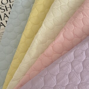 Puede incluir: Cinco tiras de tela acolchada dispuestas en diagonal en tonos pastel de azul claro, amarillo pálido, crema, rosa suave y lavanda. Cada pieza presenta un patrón de costura ondulado y redondeado constante en la superficie del material.